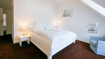 Une chambre à coucher avec un lit blanc, deux tables de chevet avec des lampes, une chaise et deux tableaux encadrés accrochés au mur.