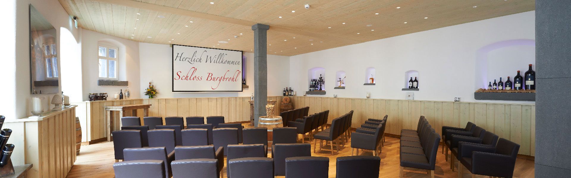 A conference room with rows of chairs, a wooden reception desk, and a wall-mounted screen displaying text.