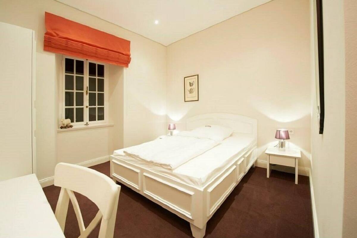 Un dormitorio con una cama individual, mesitas de noche, una ventana con cortinas, un cuadro enmarcado en la pared y una silla.