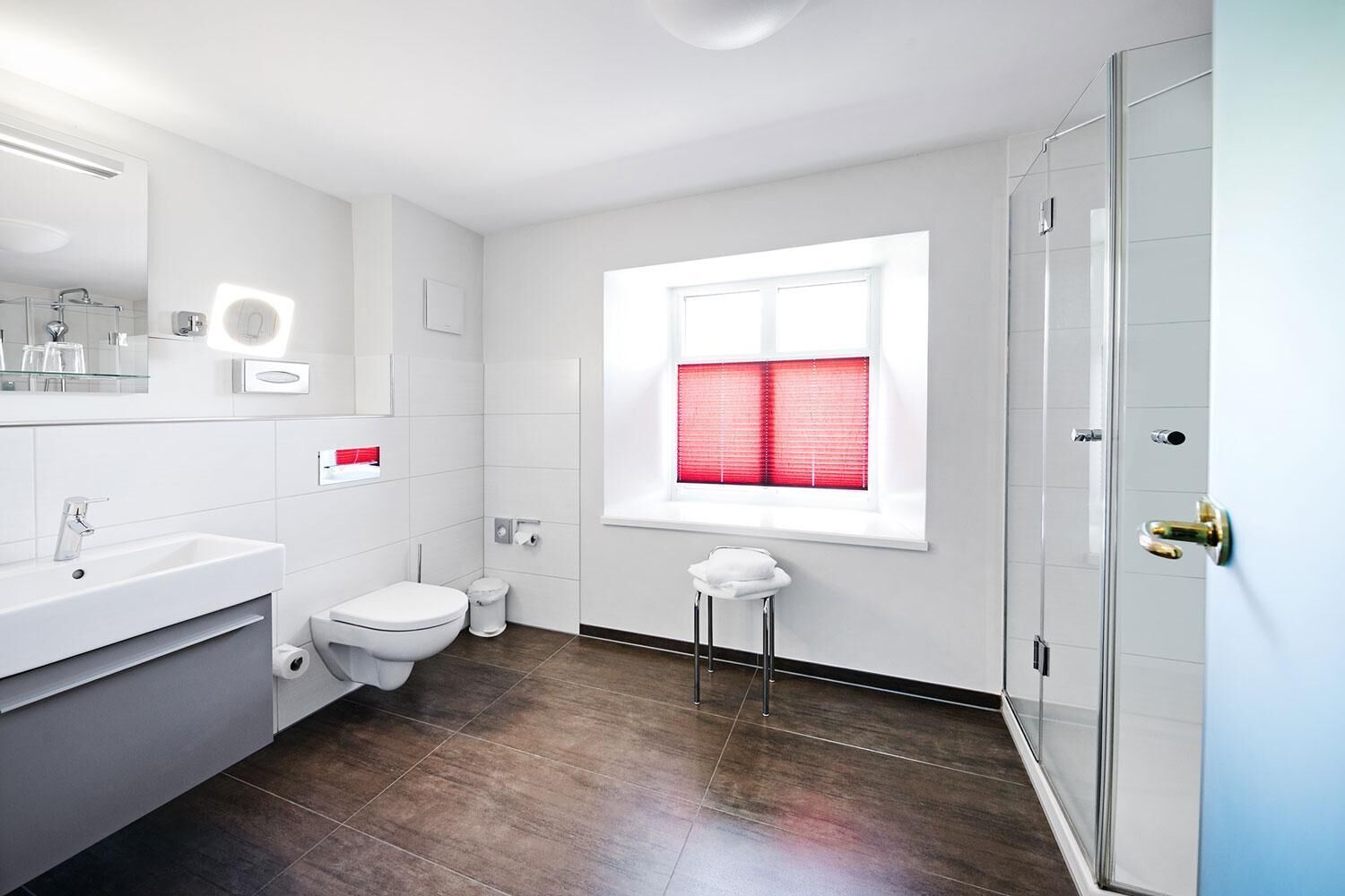 Ein modernes Badezimmer mit einem weißen Waschbecken, einer Toilette und einer Dusche. Es gibt ein Fenster mit roten Jalousien und einen kleinen Hocker.