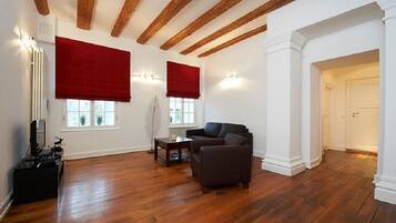 A spacious living room with hardwood flooring, a dark sofa, a coffee table, and a fireplace. There are two windows with red blinds, and a white door leading to another room.