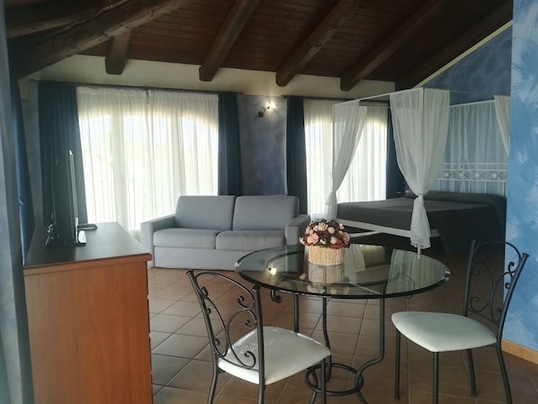 Una habitación con una mesa de vidrio, dos sillas, un sofá, una cama con cortinas blancas y una pared con un mural azul.