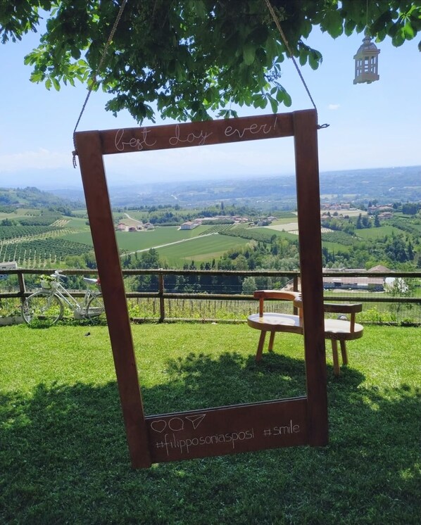 A wooden frame with the text "Best day ever!" hanging from a tree, overlooking a scenic landscape with fields and a bicycle in the foreground. - Bricco Rosso (Farigliano)