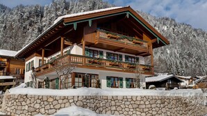 A multi-story wooden chalet with balconies, surrounded by snow-covered trees and mountains.