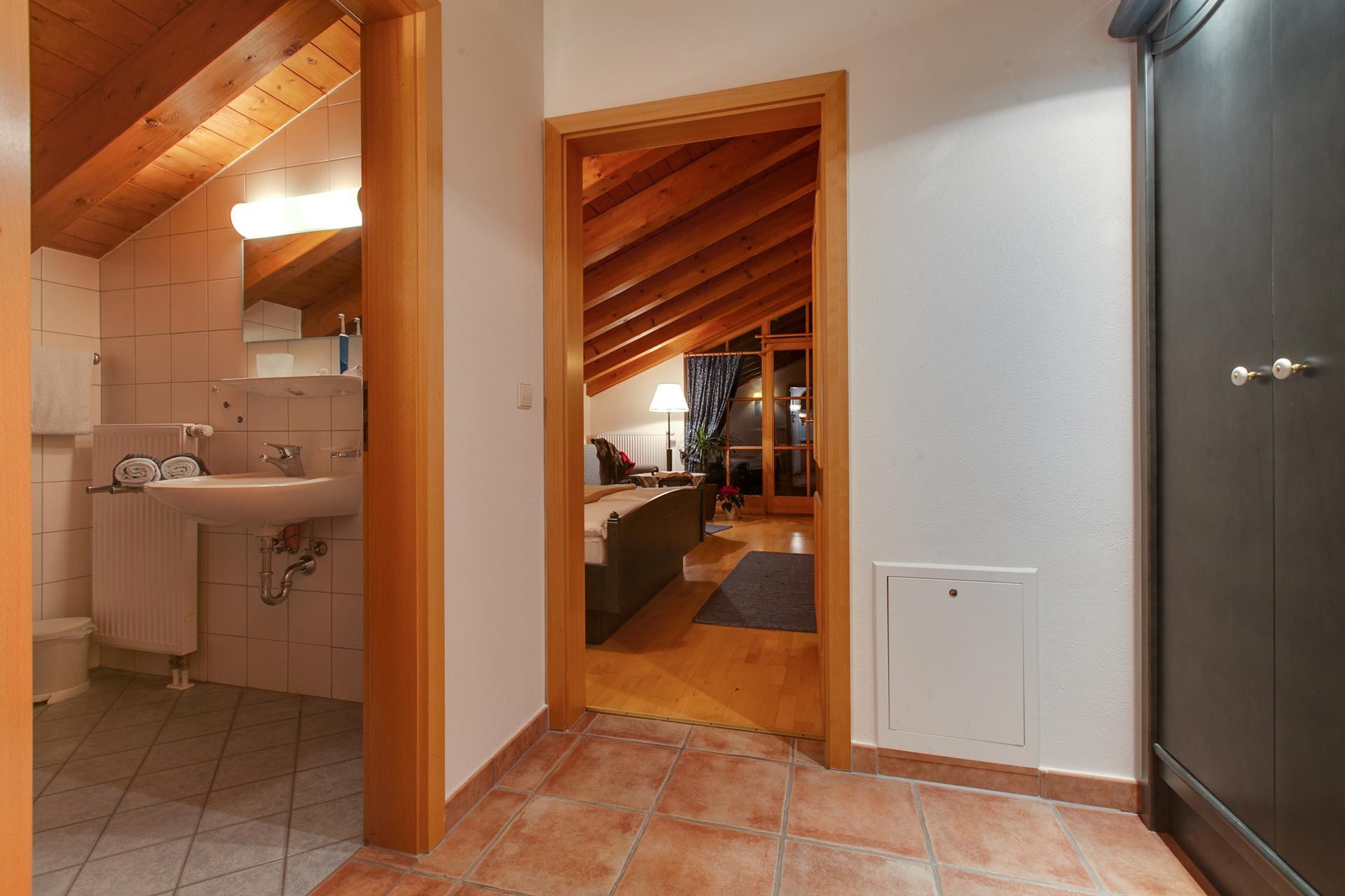 A narrow hallway with a wooden ceiling and a tiled floor leading to a bedroom with a bed, a nightstand, and a window.