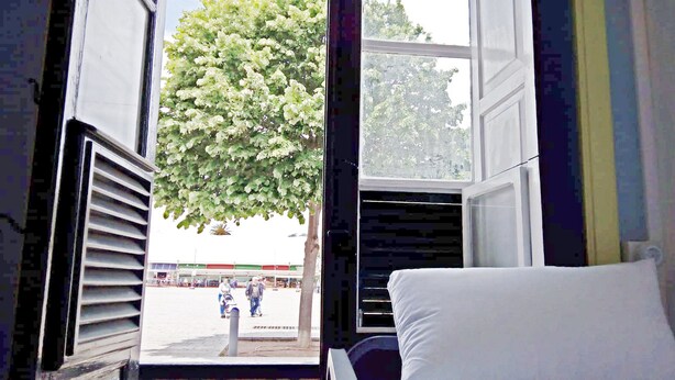 Una habitación con cama, ventana y vista a la calle, donde se ve un árbol y un edificio.