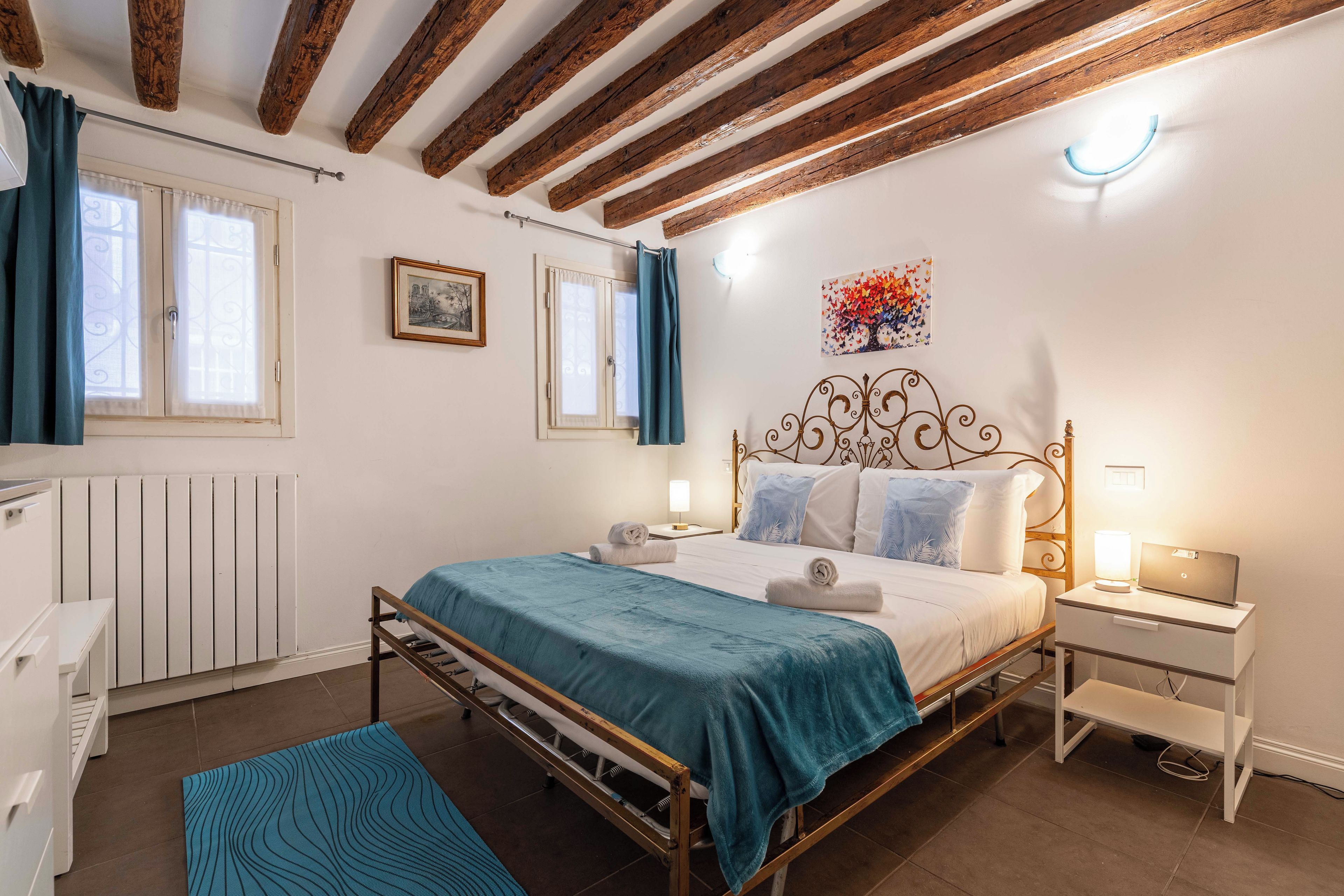 Un dormitorio con estructura de cama metálica, ropa de cama blanca, una manta azul y un radiador.