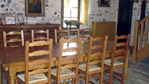 A dining room with a wooden table and chairs, a stone wall with artwork, a window with curtains, and a radiator. - Le Moulin de la Côte - Chambre d'Hôtes (Saint-Vigor)