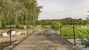 A wooden deck with white chairs and a table, surrounded by a chain-link fence and greenery. - Le Moulin de la Côte - Chambre d'Hôtes (Saint-Vigor)