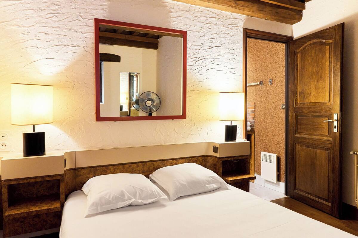 A hotel room with a bed, two bedside lamps, a mirror, a wooden wardrobe, and a wall-mounted heater.