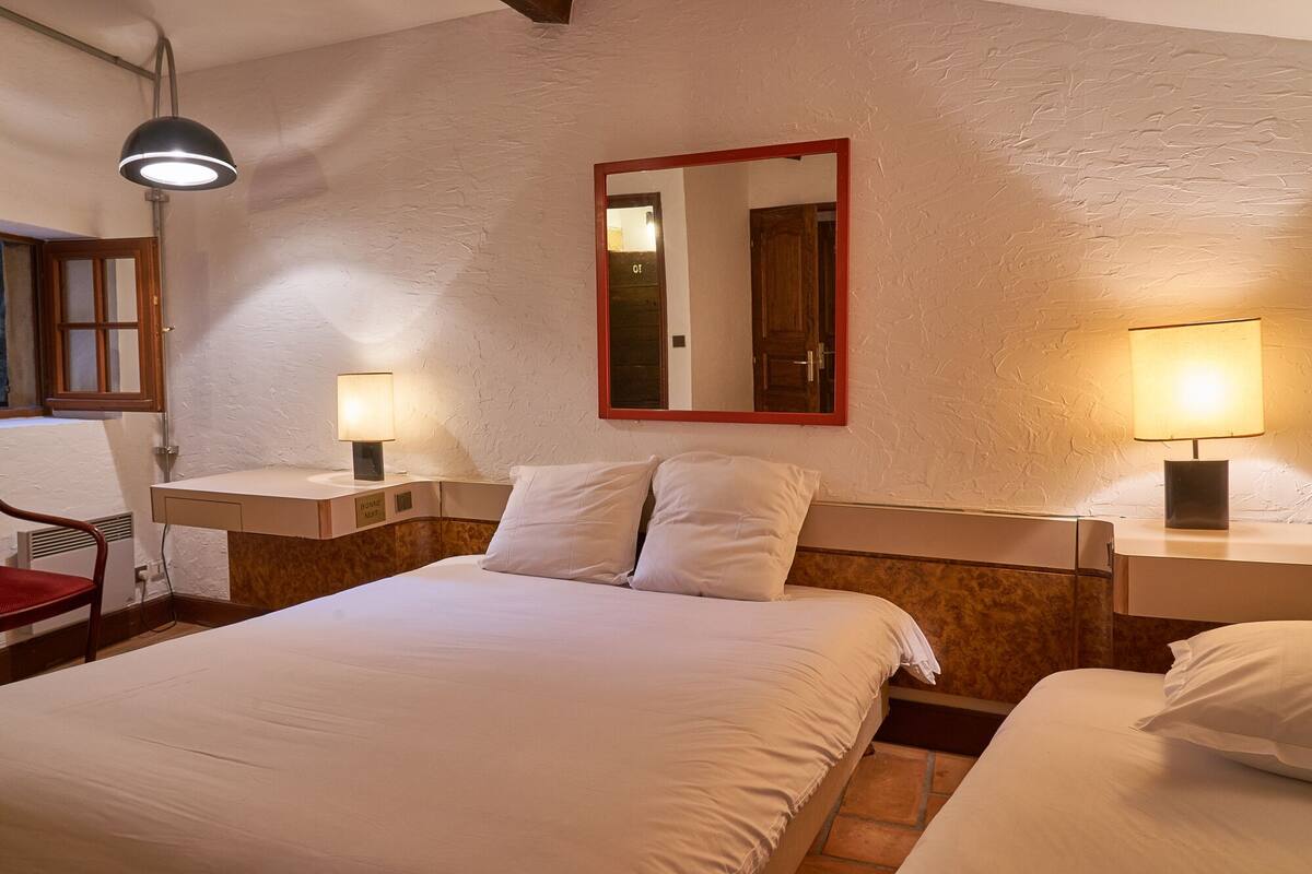 A hotel room with a large bed, two bedside tables with lamps, a mirror with a red frame, and a window with wooden shutters.