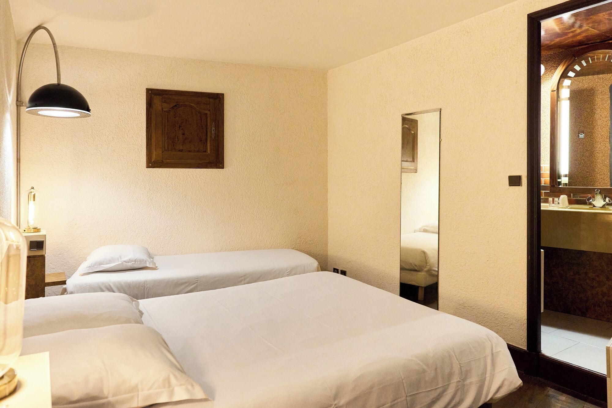 Una habitación de hotel con dos camas individuales, una puerta de madera, un espejo y una mesita de noche con lámpara.