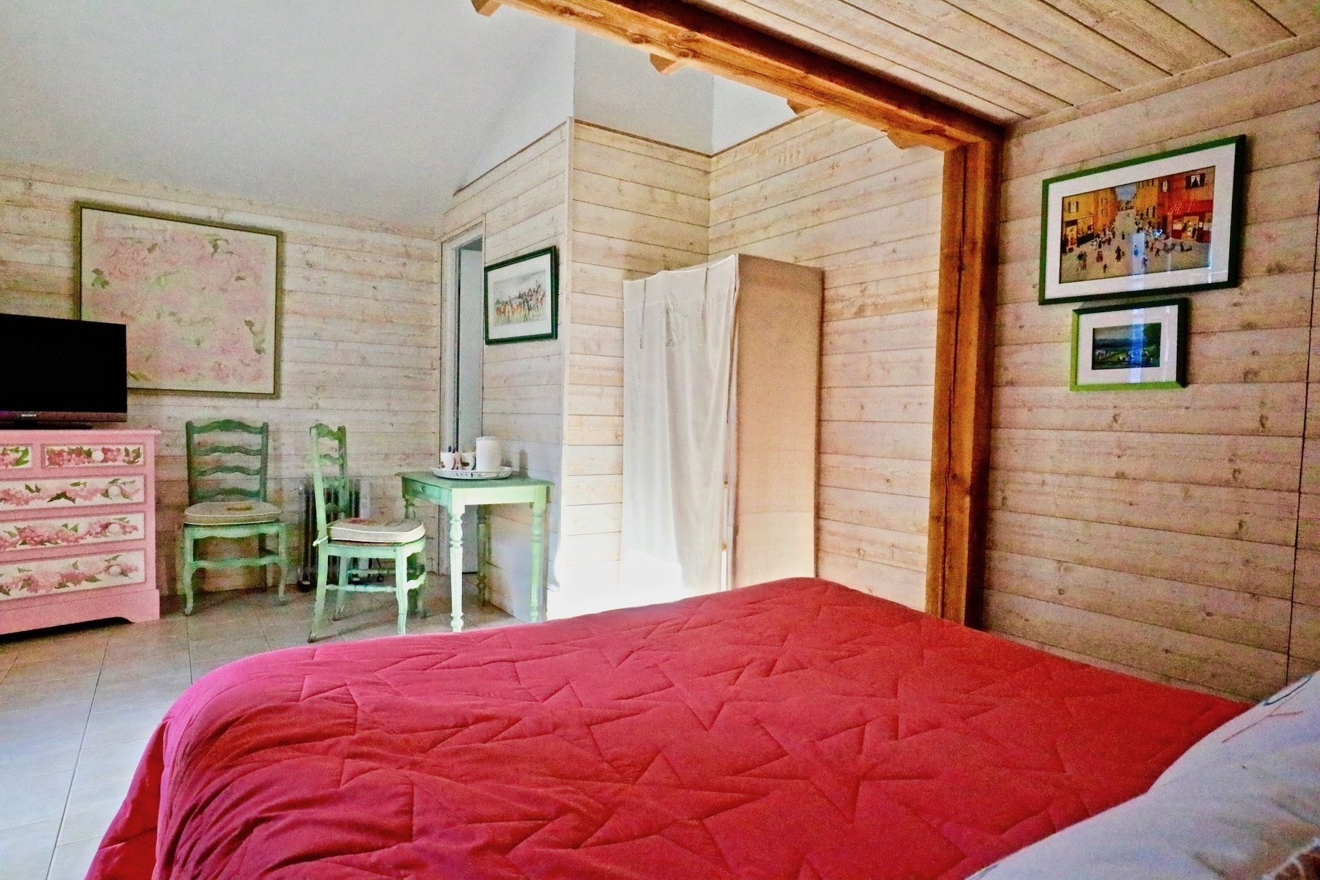 Ein Schlafzimmer mit roter gesteppter Bettdecke, Holzboden, einem hölzernen Schminktisch, einem Fernseher und zwei Stühlen.