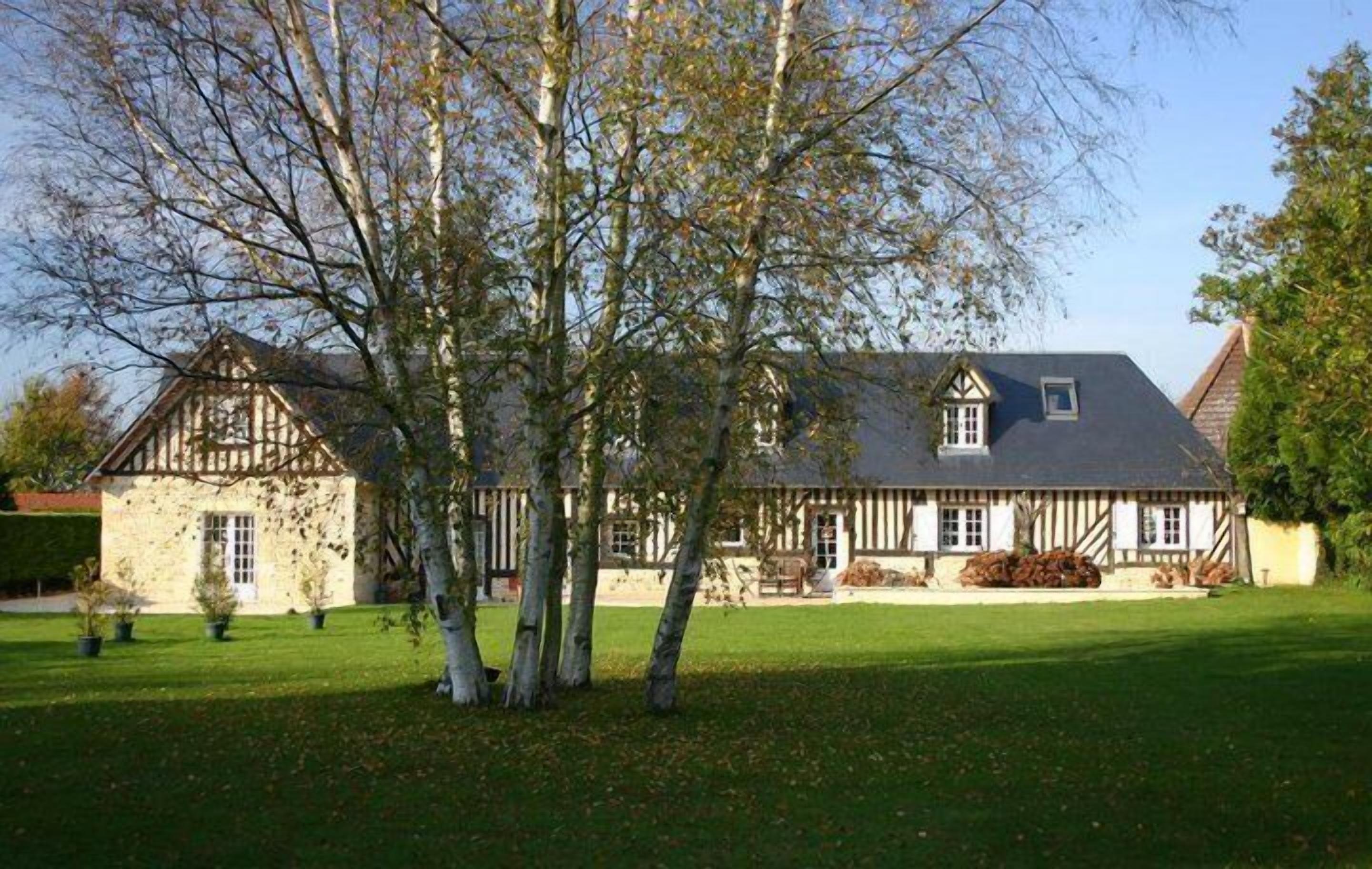 Une maison traditionnelle à colombages, dotée d’un toit en pente, entourée d’une pelouse bien entretenue et de grands arbres.