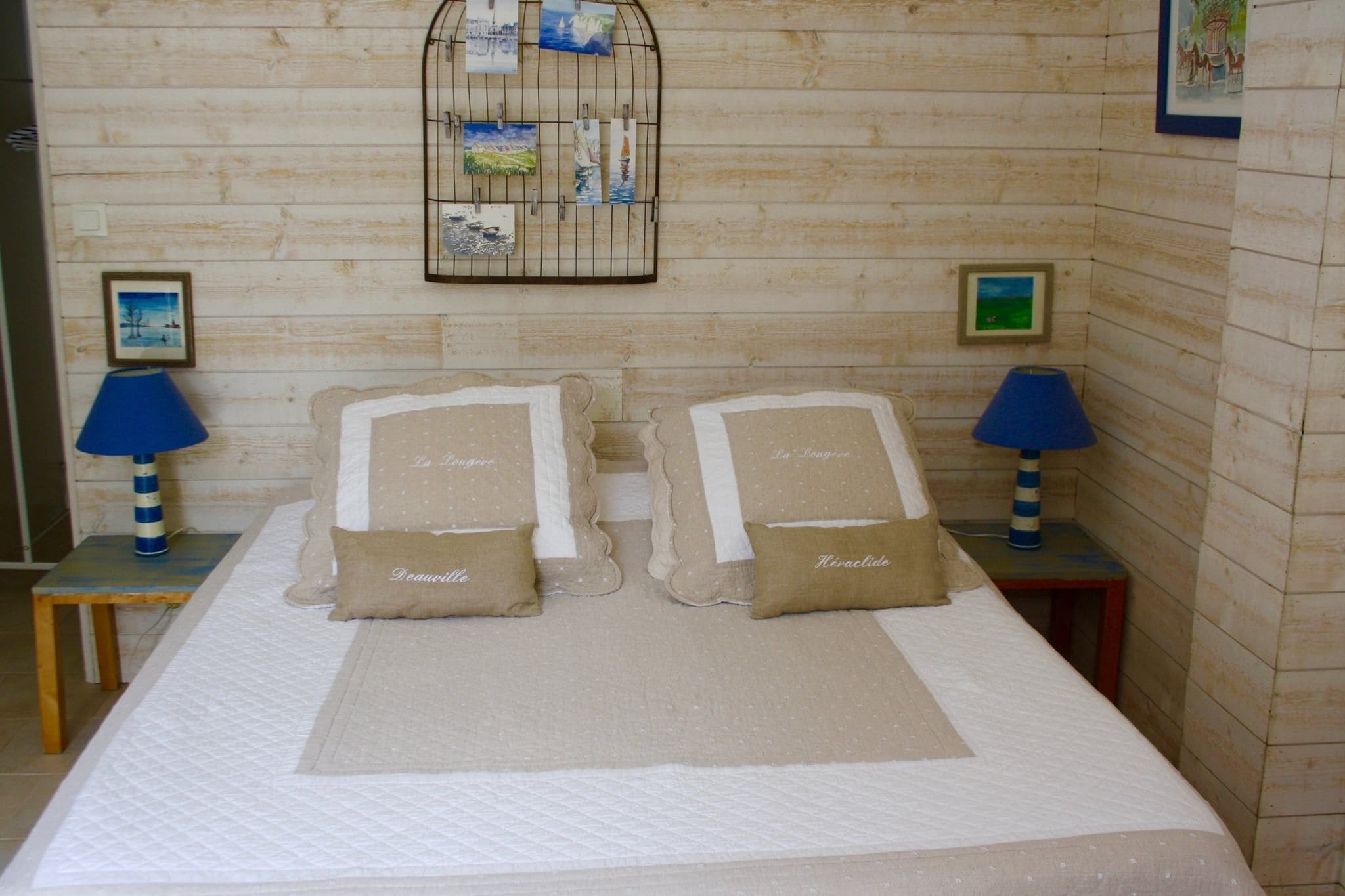 Una cama impecablemente hecha con dos almohadas etiquetadas "Le Trou" y "Le Trou" en una habitación con paredes de madera, una pequeña mesa con una lámpara y un expositor de fotos en la pared.