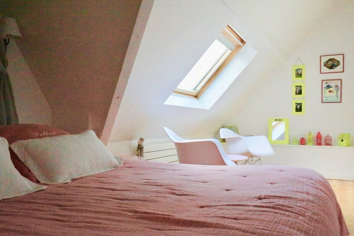Un dormitorio con techo inclinado, una cama con ropa de cama rosa, una silla blanca, un escritorio con espejo y un tragaluz.