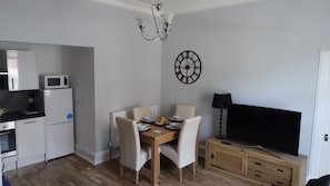 A compact kitchen with white cabinets, a microwave, and a refrigerator. A dining table with chairs and a clock on the wall is in the center.