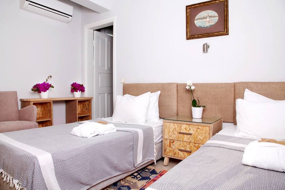 Ein Hotelzimmer mit zwei Betten, einem Holz-Nachttisch, einem kleinen Tisch mit Blumenvase und einem gerahmten Bild an der Wand.