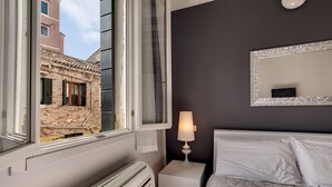 A bedroom with a bed, a nightstand with a lamp, a window with a view of a building, and a radiator. - Charming Castello Apartments by Wonderful Italy (Venice)