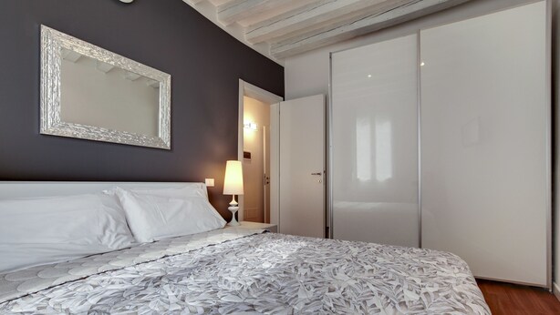 A bedroom with a large bed, a mirror, a wardrobe, and a nightstand with a lamp. - Charming Castello Apartments by Wonderful Italy (Venice)