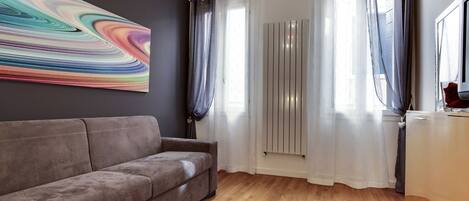 A modern living room with a grey sofa, a colorful abstract painting, and a large window with sheer curtains.