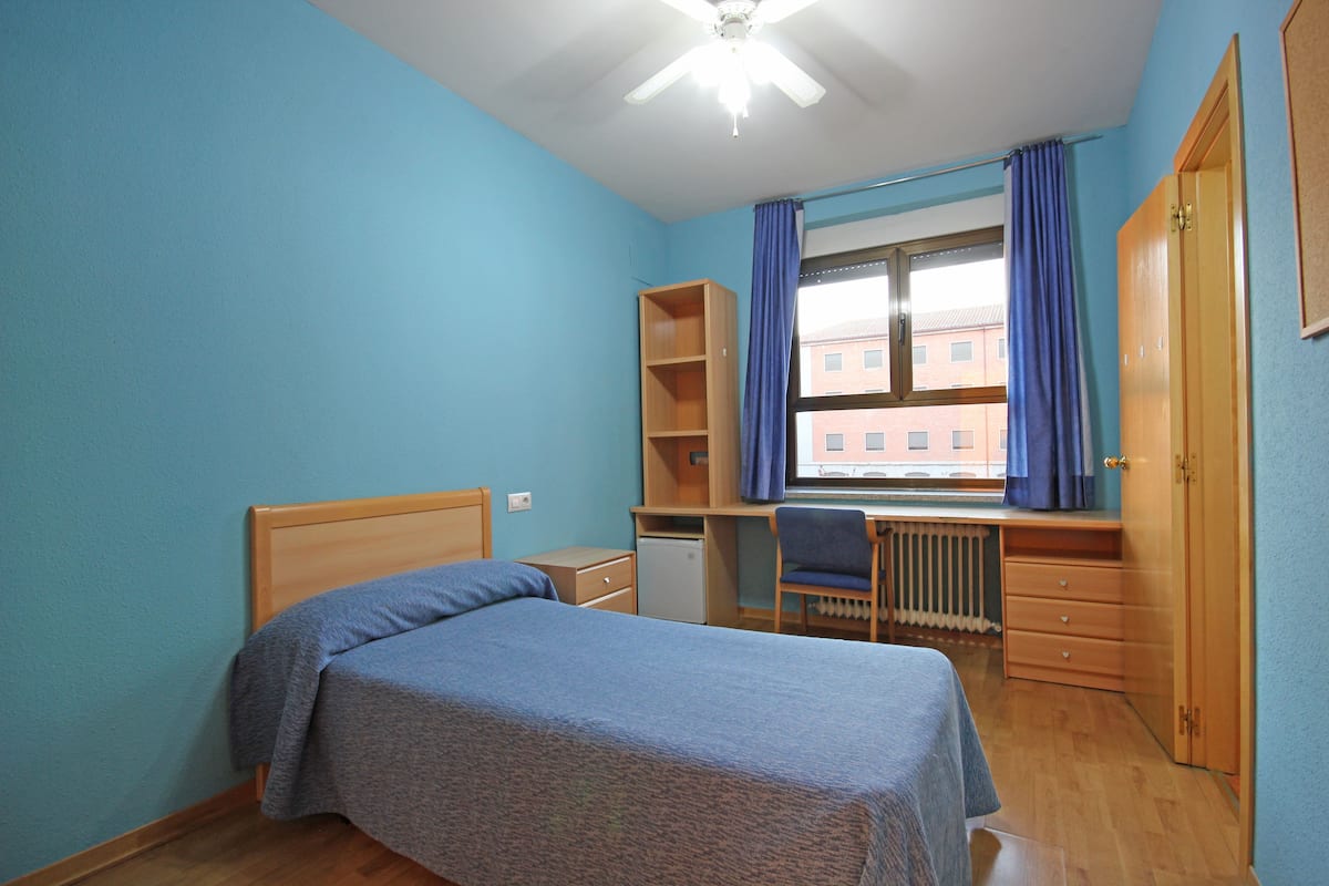 A bedroom with a bed, a desk, a chair, a radiator, and a window with curtains.