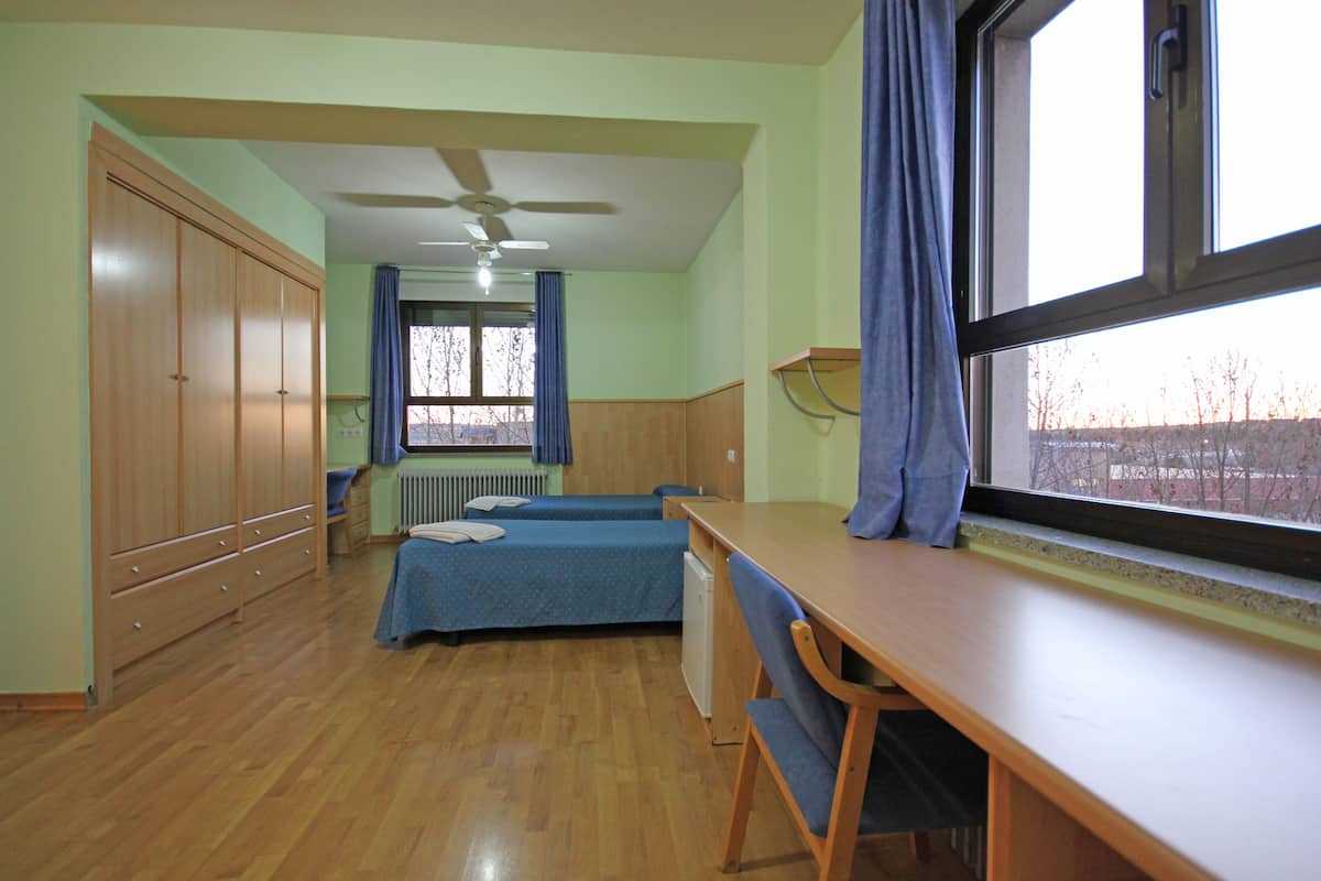 A dormitory room with two beds, a desk, a chair, a radiator, and a window with curtains.