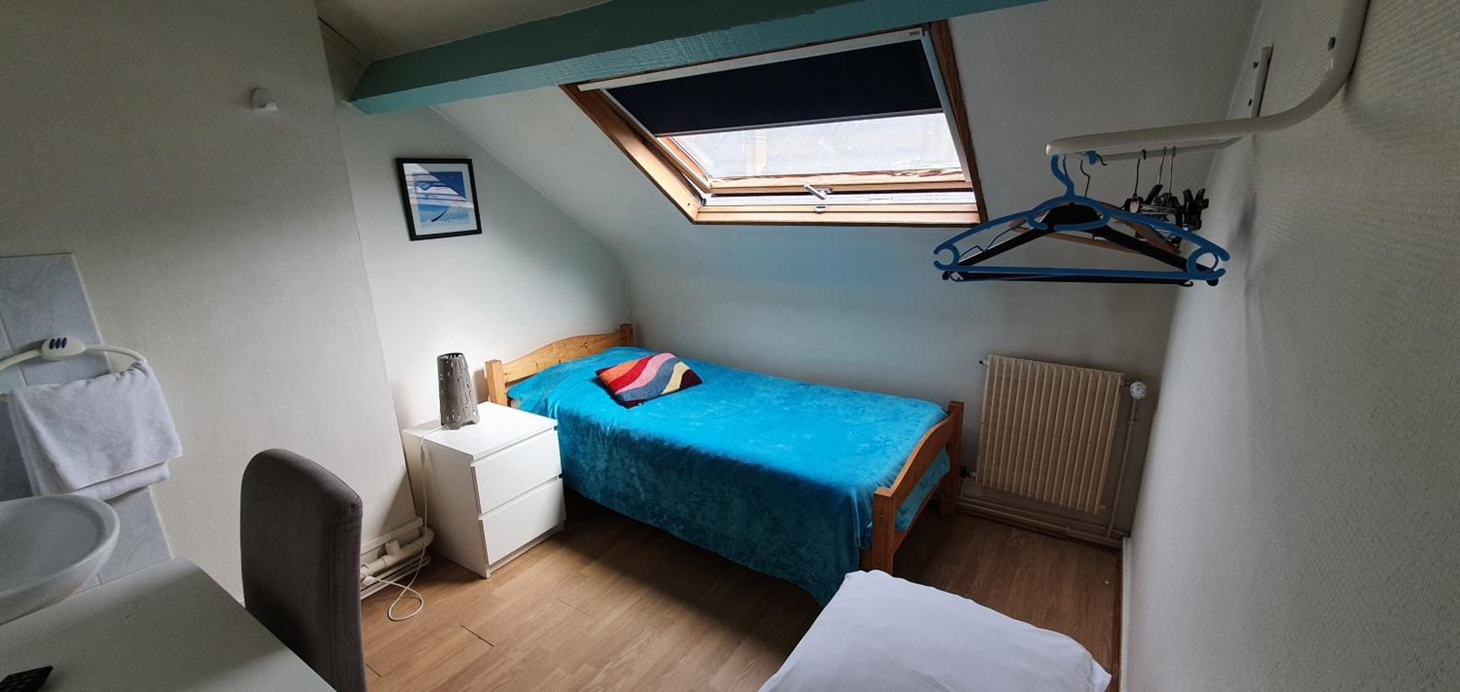 A compact bedroom with a single bed, a small bedside table, a chair, a radiator, and a skylight.