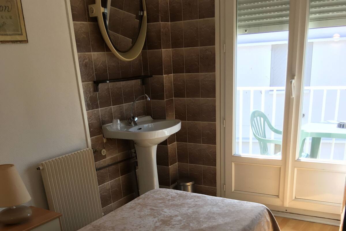 Une chambre avec un lit, une table de chevet avec une lampe, un radiateur, une salle de bain avec un lavabo et un miroir, et un balcon avec une chaise.