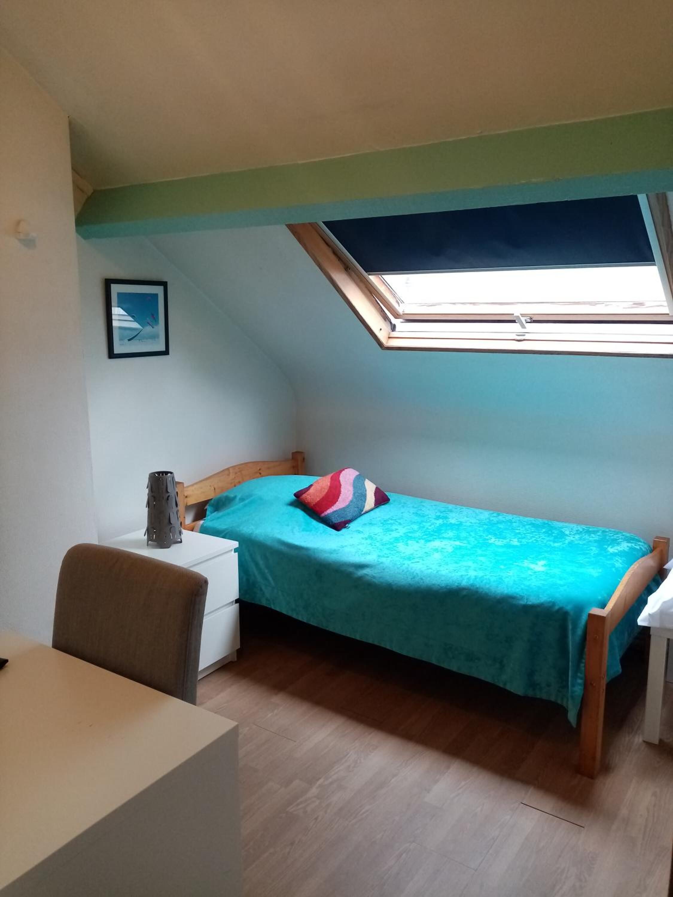 Une petite chambre avec un lit simple, une lucarne, une table de chevet, une chaise et un bureau.