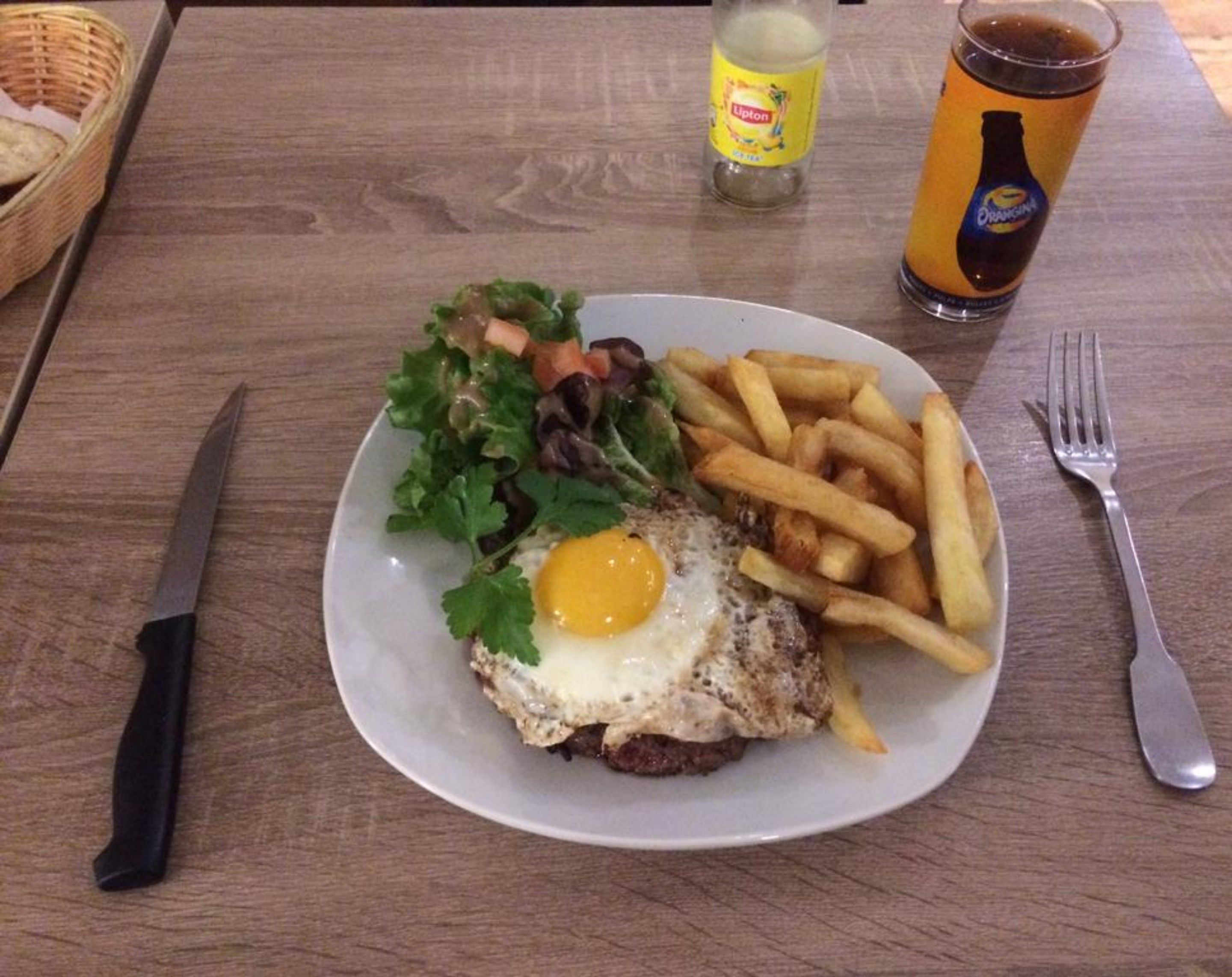 A plate with a burger, egg, fries, and salad, a bottle of Lipton tea, and a glass of beer on a wooden table.
