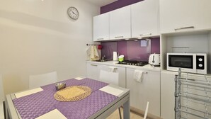 A compact kitchen with white cabinets, a microwave, and a stove. There is a clock on the wall and a table with a purple tablecloth. - Venetian Dream in Biennale Area by Wonderful Italy (Venice)
