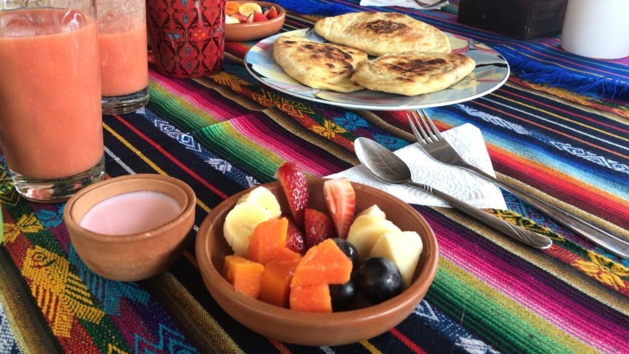 A colorful woven tablecloth, a bowl of fruit, a plate of flatbreads, and two glasses of a pinkish beverage.