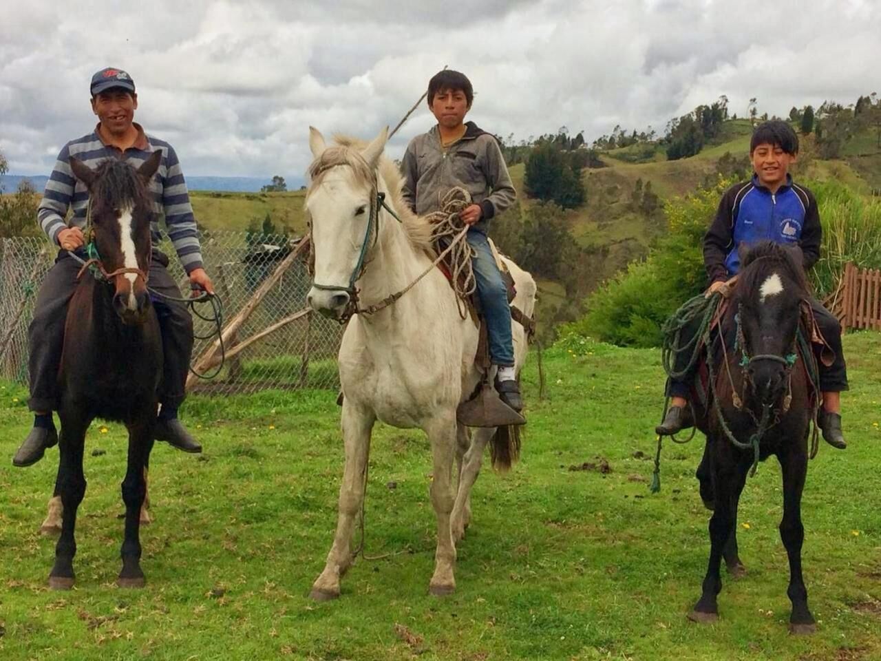 Three individuals on horses in an outdoor setting with grassy terrain and a fence in the background.