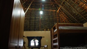 Una habitación con techo de paja, una cama litera, una mesita con silla y una ventana con cortinas.