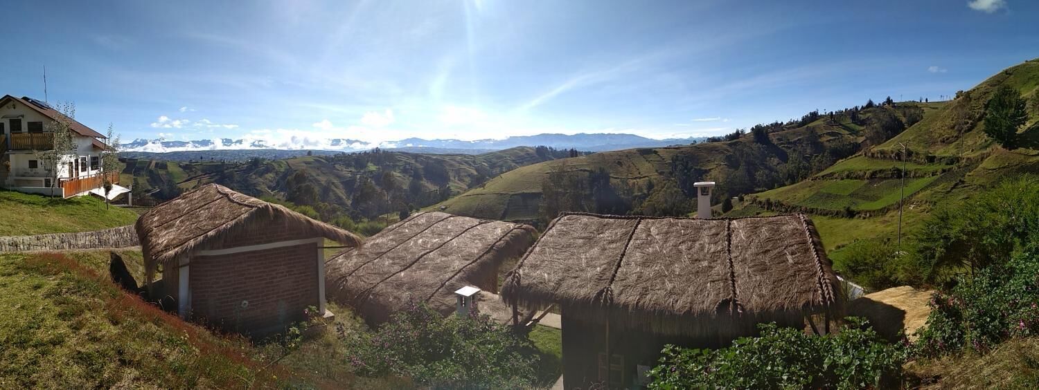 Paisaje rural con casas tradicionales de techo de paja, un edificio de ladrillo y vistas a montañas distantes.