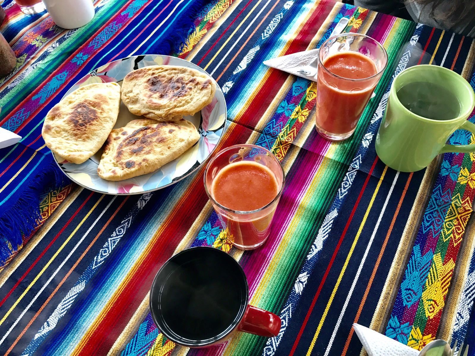 Un mantel tejido de colores con un plato de panes planos, dos vasos de bebida roja y una taza verde.