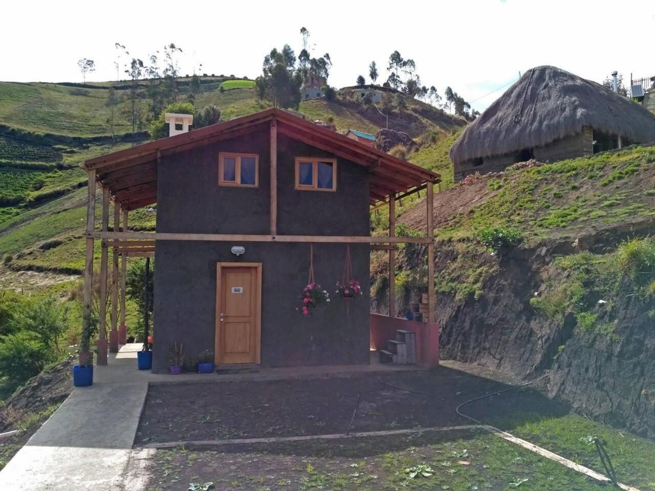 Un edificio de dos pisos con puerta y ventanas de madera, rodeado de follaje y colinas.