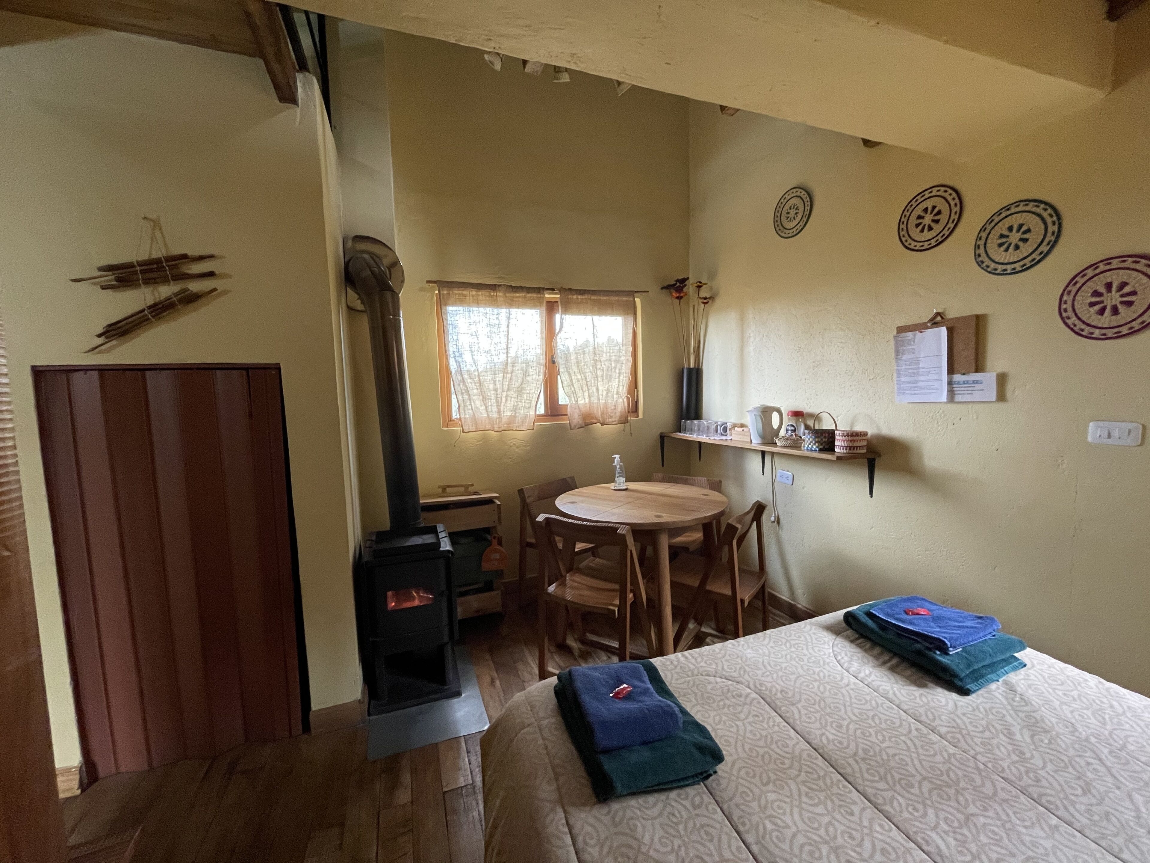 A cozy room with a wooden floor, a bed with a patterned bedspread, a small round table with chairs, a window with curtains, and a wall-mounted shelf with various items.