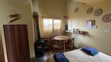 A cozy room with a wooden floor, a bed with a patterned bedspread, a small round table with chairs, a window with curtains, and a wall-mounted shelf with various items.