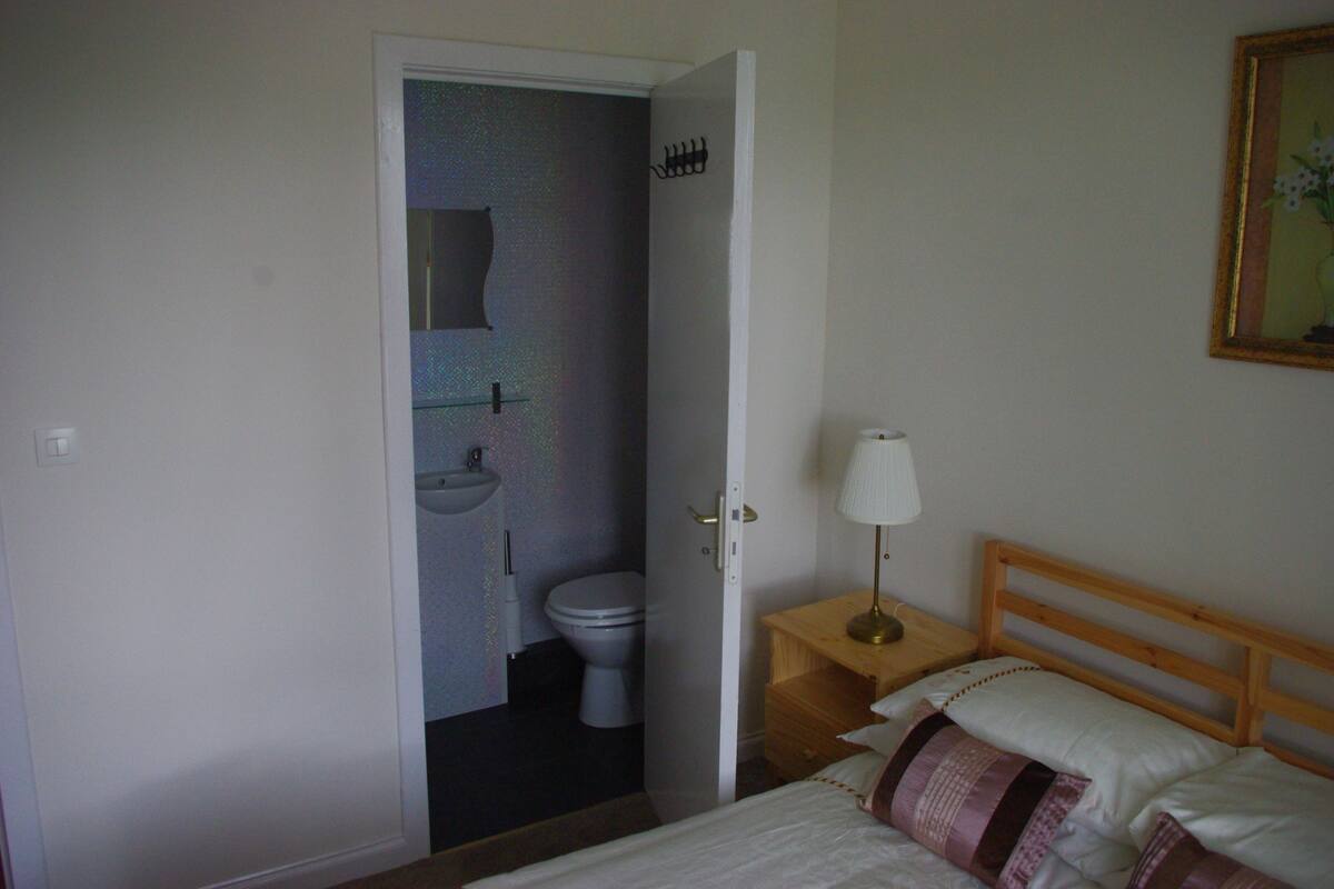 A bedroom with a bed, a bedside table with a lamp, a wooden headboard, and a door leading to a bathroom with a toilet and sink.