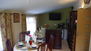 A dining area with a table set for four, chairs, a television mounted on the wall, and a kitchen area with cabinets and appliances. - La Maison Celtique Chambres d'hôtes (Saint-Nicolas-du-Pelem)