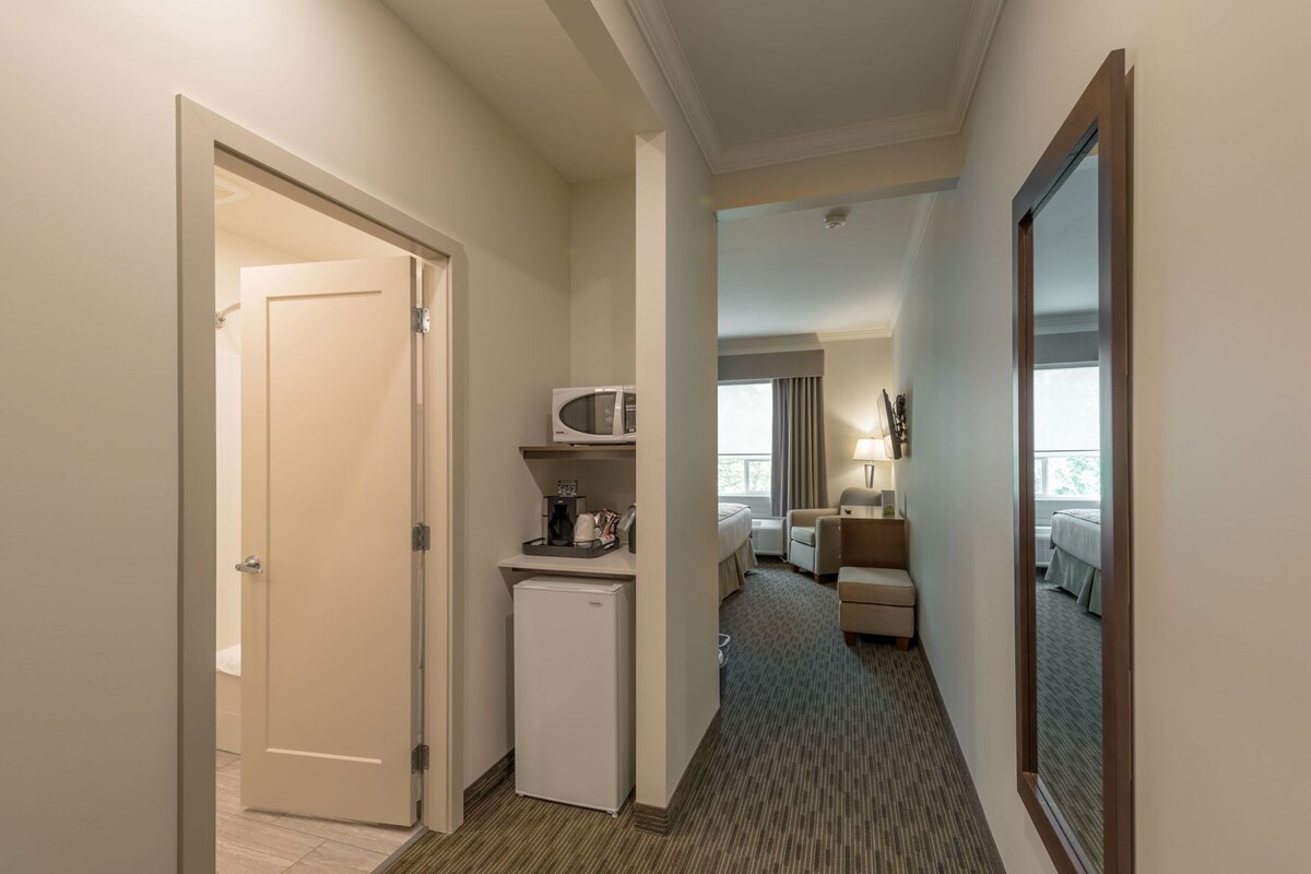 A hotel hallway with a door, a microwave, a refrigerator, a bed, a chair, and a lamp.