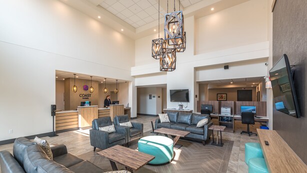 A spacious lobby with leather sofas, wooden tables, and a central ottoman. A reception desk is visible in the background with a sign reading "COAST hotels." - Coast Oliver Hotel (Oliver)