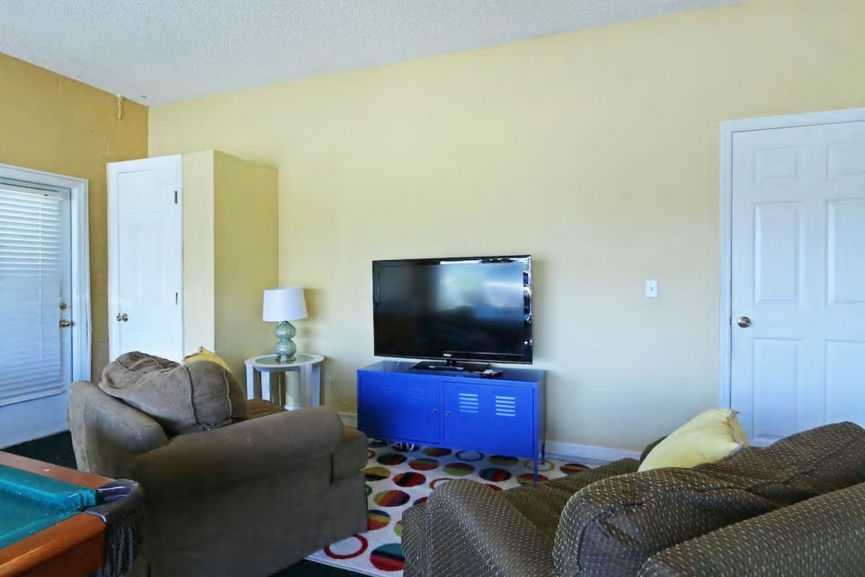 Ein Wohnzimmer mit Fernseher auf einem blauen Schrank, zwei Sofas, einem kleinen Tisch mit Lampe und einem Billardtisch.
