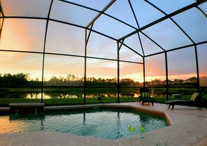 A glass-enclosed swimming pool with a view of a sunset over a lake, surrounded by chairs and a small garden area.