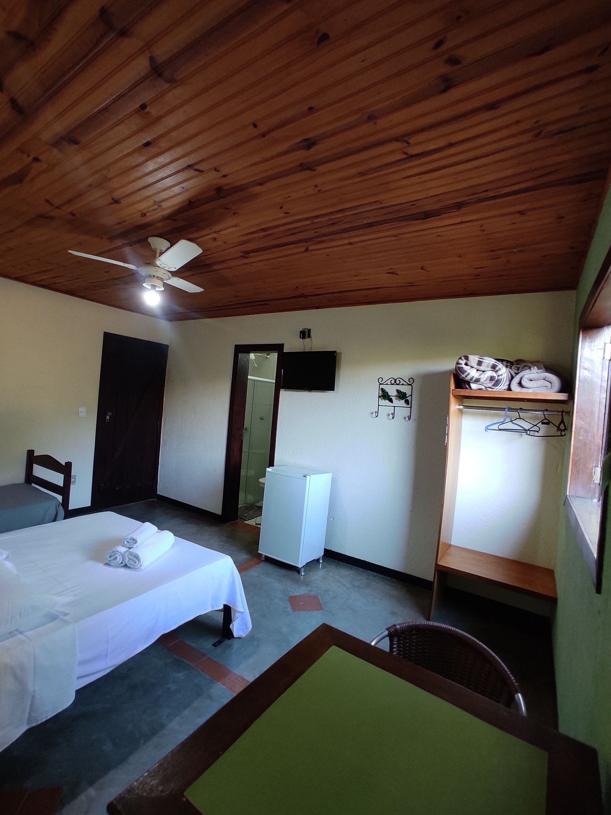 A room with a bed, a chair, a small table, a ceiling fan, a TV, and a refrigerator.