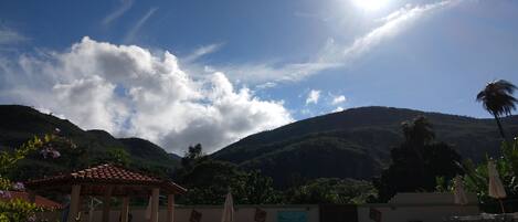 A swimming pool with a tiled edge, surrounded by lounge chairs and umbrellas, set against a backdrop of mountains and a clear sky.