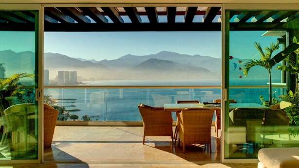 A modern outdoor patio with a glass balustrade, wicker furniture, and a view of the ocean and mountains. - Ph Toshi (Puerto Vallarta)