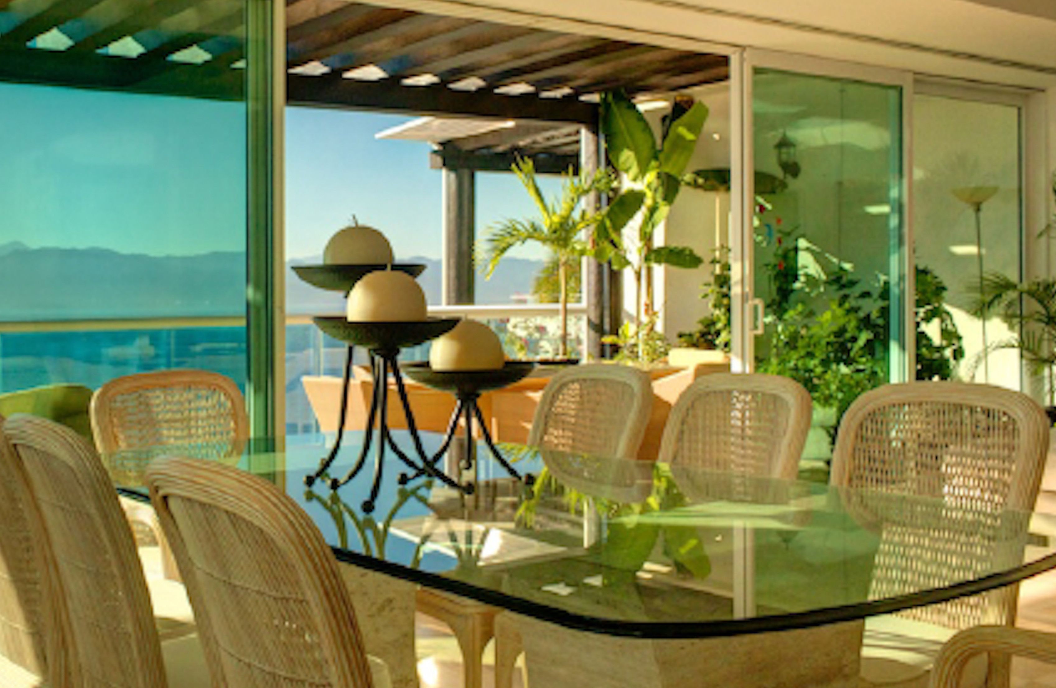 Una mesa de comedor con cubierta de vidrio, sillas de mimbre, barandilla de cristal y vista al océano.